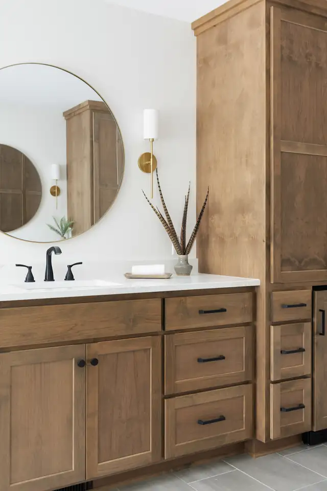 Bathroom remodel with natural wood vanity and tall linen cabinet, round mirror and brass sconce