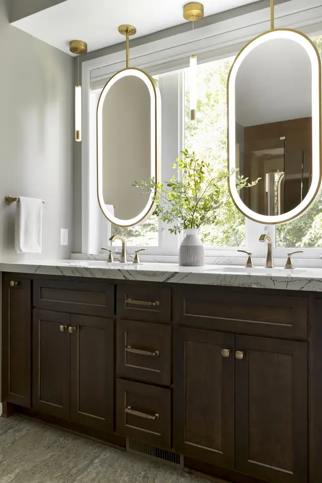 Lakeville primary bath with a dark wood vanity, two oval lighted mirrors and brass faucets